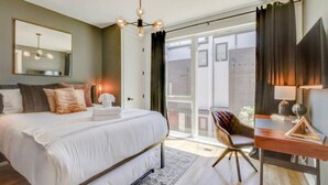 A modern bedroom with a large bed, a desk, and a chair. There is a large window with curtains and a view of the outside. - Midtown Skyline Retreat | Rooftop Deck | Game Room (Nashville)