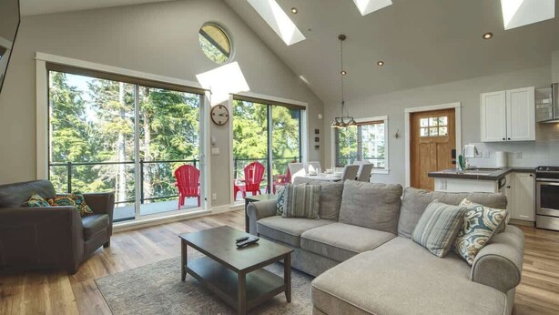 Ein geräumiges Wohnzimmer mit einer großen Couch, einem Couchtisch und einem Essbereich mit roten Stühlen. Dachfenster und große Fenster sorgen für natürliches Licht.