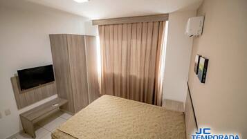 A hotel room with a bed, a TV on a wooden stand, a small bench, and a window with curtains.