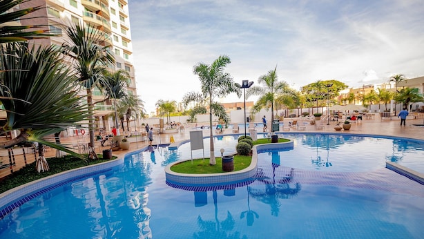A large outdoor swimming pool with multiple sections, surrounded by palm trees and lounge chairs, set against a backdrop of a high-rise building. - RIVIERA PARK - JC TEMPORADA (Caldas Novas)
