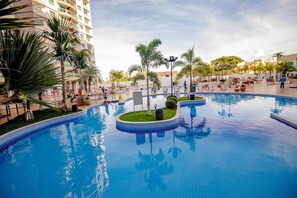 A large outdoor swimming pool with multiple sections, surrounded by palm trees and lounge chairs, set against a backdrop of a high-rise building. - RIVIERA PARK - JC TEMPORADA (Caldas Novas)