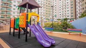 A playground with slides and climbing structures, surrounded by a plaza with benches and a building in the background. - RIVIERA PARK - JC TEMPORADA (Caldas Novas)