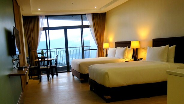 A hotel room with two beds, a desk, a chair, and a TV. There is a balcony with curtains and a view of the sky. - Alvea Hotel Baguio  (Baguio)