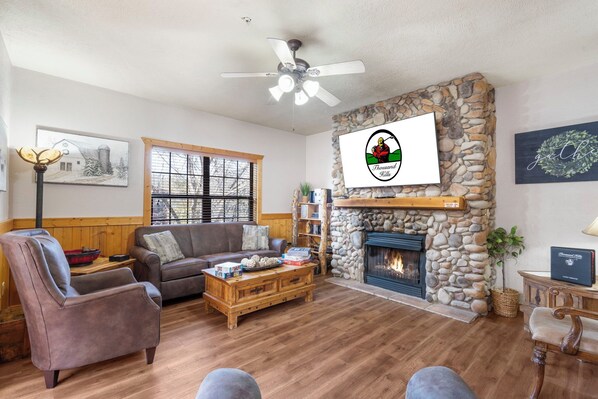 A cozy living room with a stone fireplace, a ceiling fan, and a large window. - Great Cabin, Close to the Pool, Relaxing Views! 2 Minutes to Tanger Outlets! (Branson)