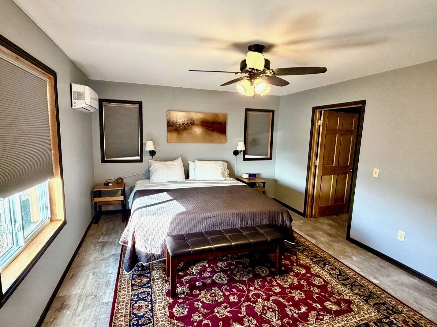 A bedroom with a bed, a bench, a ceiling fan, a nightstand, a lamp, a window with blinds, and a door.
