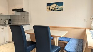 A small kitchenette with a coffee maker, a microwave, and a stove hood. There is a dining table with two chairs and a bench with a patterned cushion. A painting of a mountain landscape is on the wall. - Bergfreude 06 by Interhome (Saas-Fee)