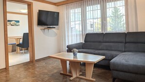 A living room with a sofa, a TV mounted on the wall, a wooden table, and a window with curtains. - Bergfreude 06 by Interhome (Saas-Fee)