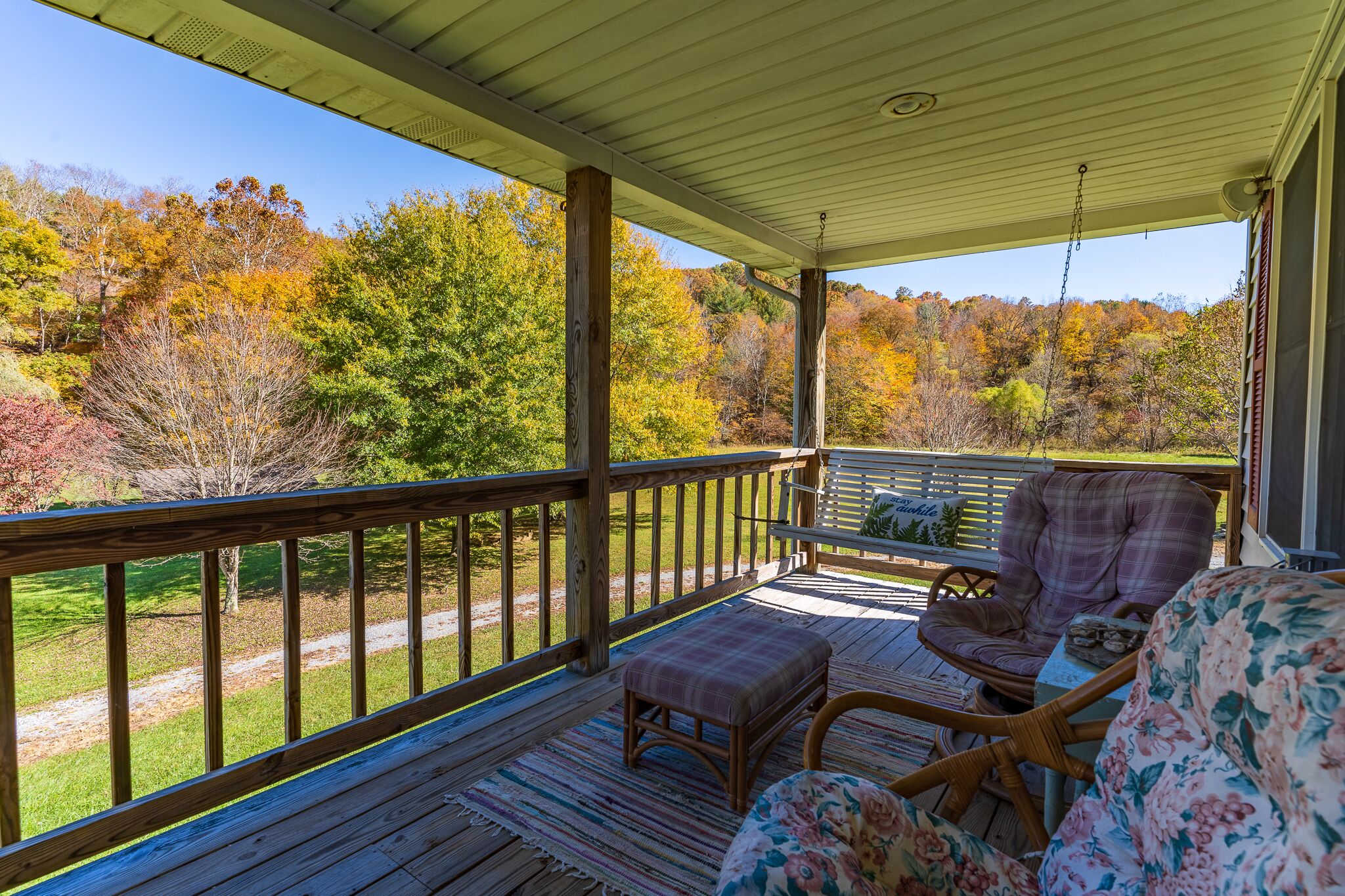 A covered porch with a wooden railing, wicker furniture, and a view of a forested area.