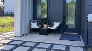 Un bâtiment moderne à l’ext érieur sombre, doté d’un balcon avec deux fauteuils en osier et une petite table, ainsi que d’une zone d’entrée pavée.