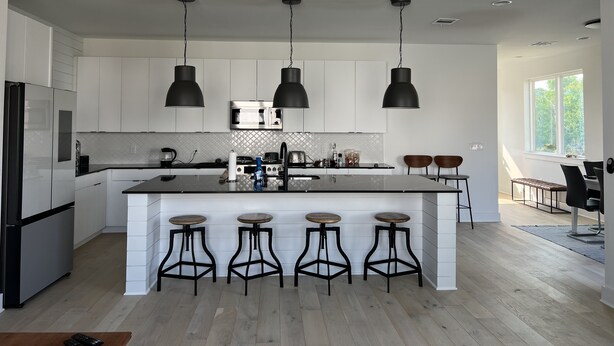 Une cuisine moderne avec des meubles blancs, un îlot central doté de tabourets, des suspensions noires et une grande fenêtre.