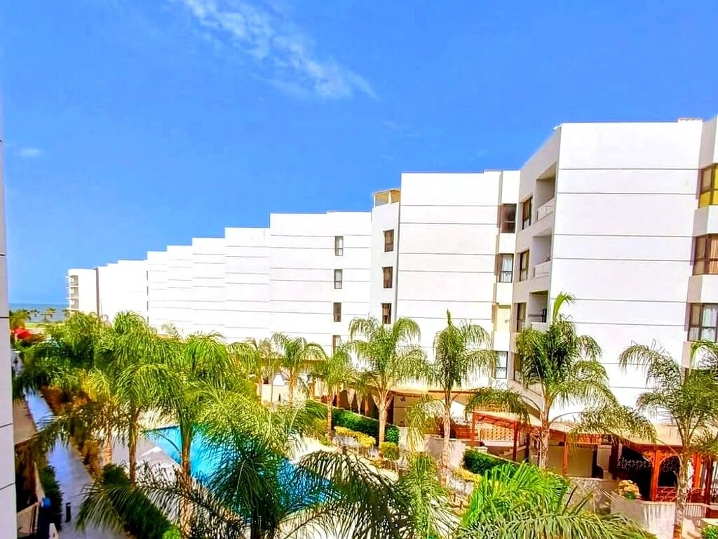 A modern hotel with a series of white buildings, a pool area with palm trees, and a clear blue sky.