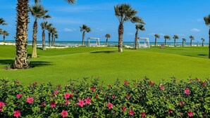 A well-maintained golf course with a clear view of the ocean, surrounded by palm trees and vibrant pink flowers. - Beachfront Chalet in Porto Said with Pool & Ga (Port Said Governorate)