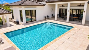 Ein rechteckiger Pool mit Fliesenveranda, umgeben von einer überdachten Terrasse mit Stühlen und Tischen.