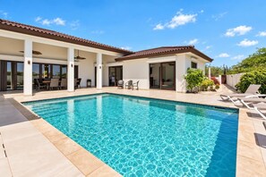 A swimming pool with a tiled deck, a covered patio area with chairs, and a view of the surrounding landscape.