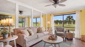 A spacious living room with a large window, a ceiling fan, and a round coffee table.