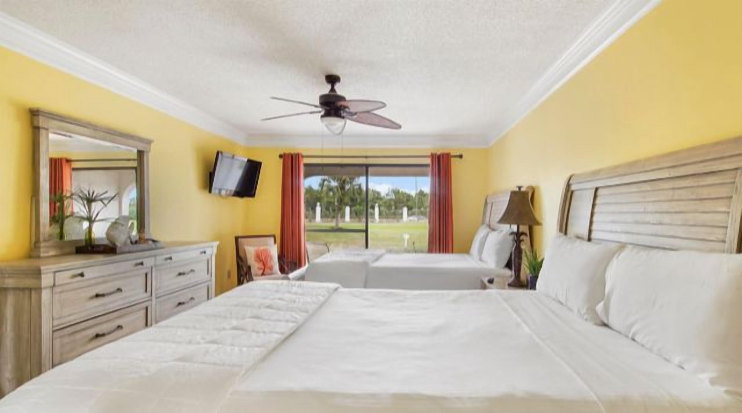 A hotel room with two beds, a ceiling fan, a TV, and a view of the outdoors.