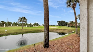 A golf course with a pond, palm trees, and a building in the background. - Edgewater Villa 1704, Top Resort, Beach Tram, Gated sleeps 4 (Panama City Beach)
