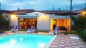 Una piscina con sillones de descanso y sombrillas, un área de patio y un edificio con techo de tejas.