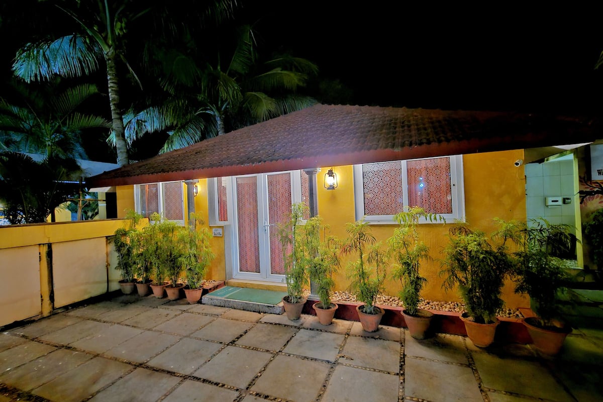 Un edificio amarillo con techo de tejas y múltiples ventanas con diseños decorativos. Varias plantas en macetas están dispuestas a lo largo de la fachada.