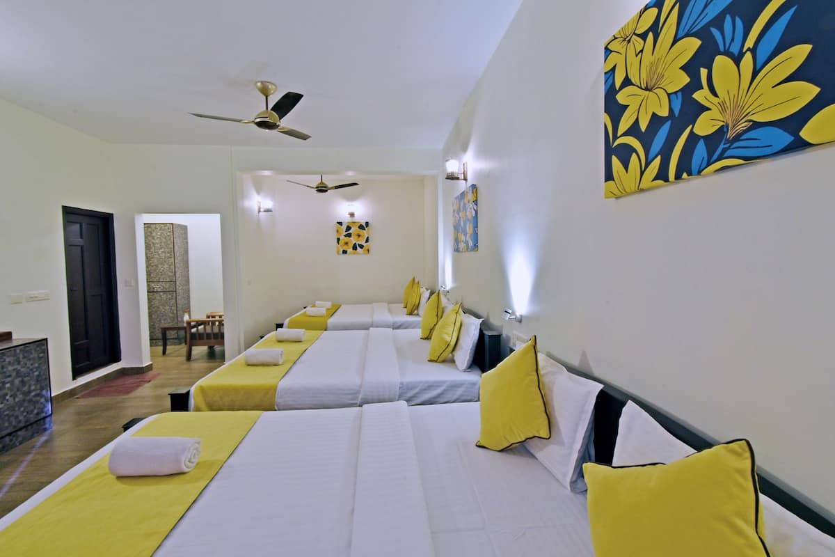 Habitación de hotel con tres camas, cada una con ropa blanca y almohadas amarillas. Cuenta con ventiladores de techo, luces fijadas a la pared y obras de arte en las paredes.