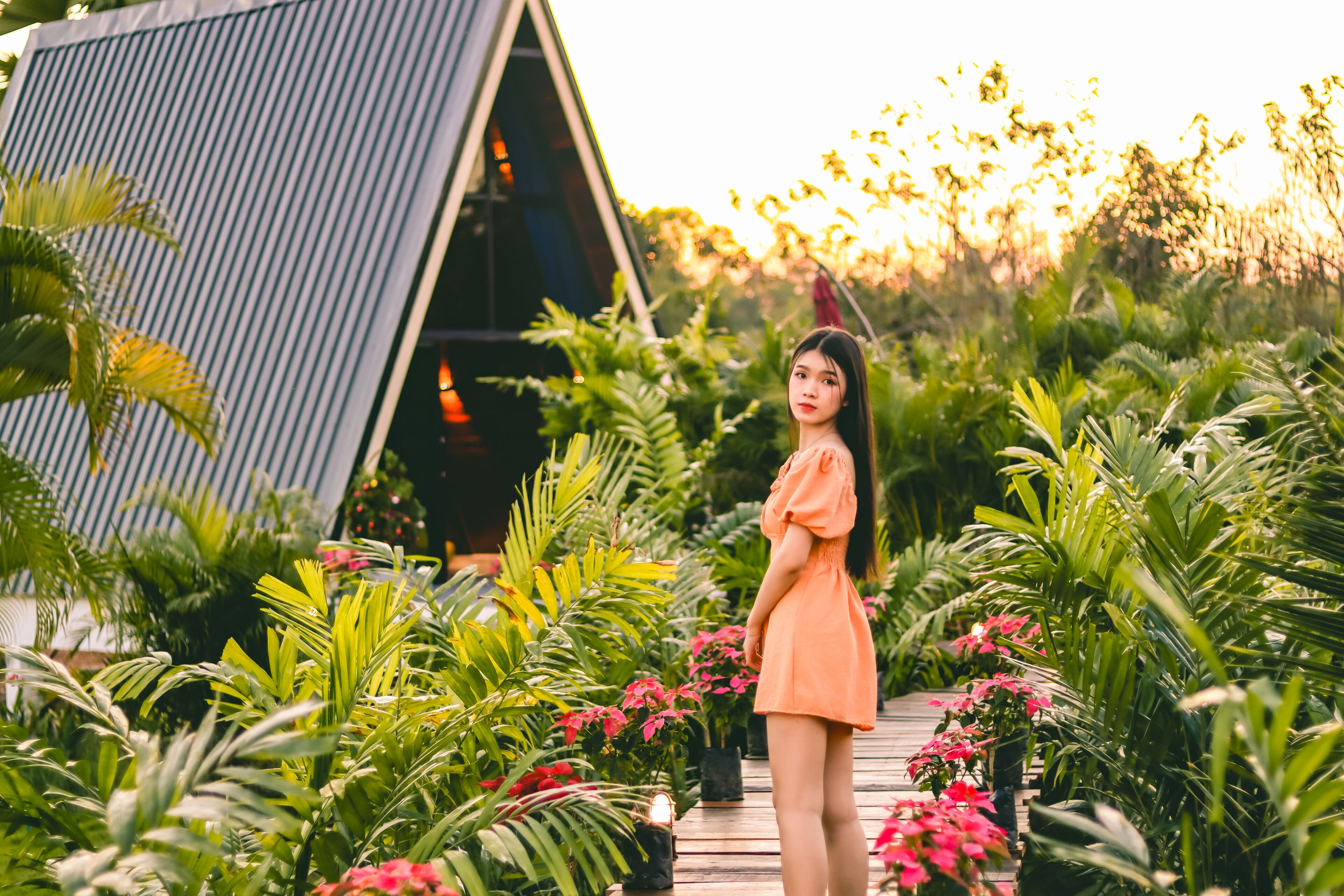 A woman in an orange dress standing on a wooden deck surrounded by lush greenery and a modern building structure.