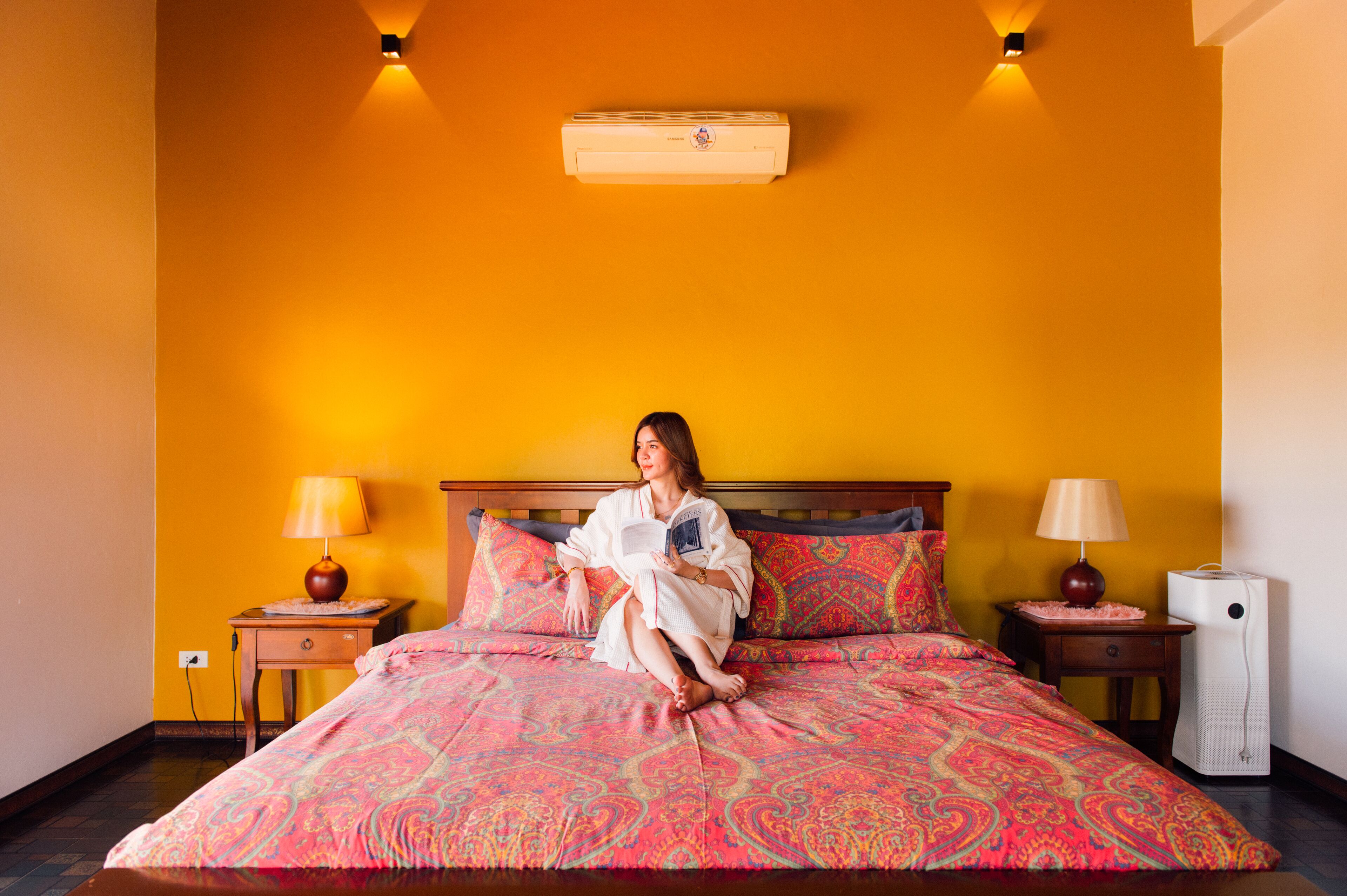 A bed with a patterned bedspread, two bedside lamps, a nightstand, a wall-mounted air conditioner, and a wall sconce.