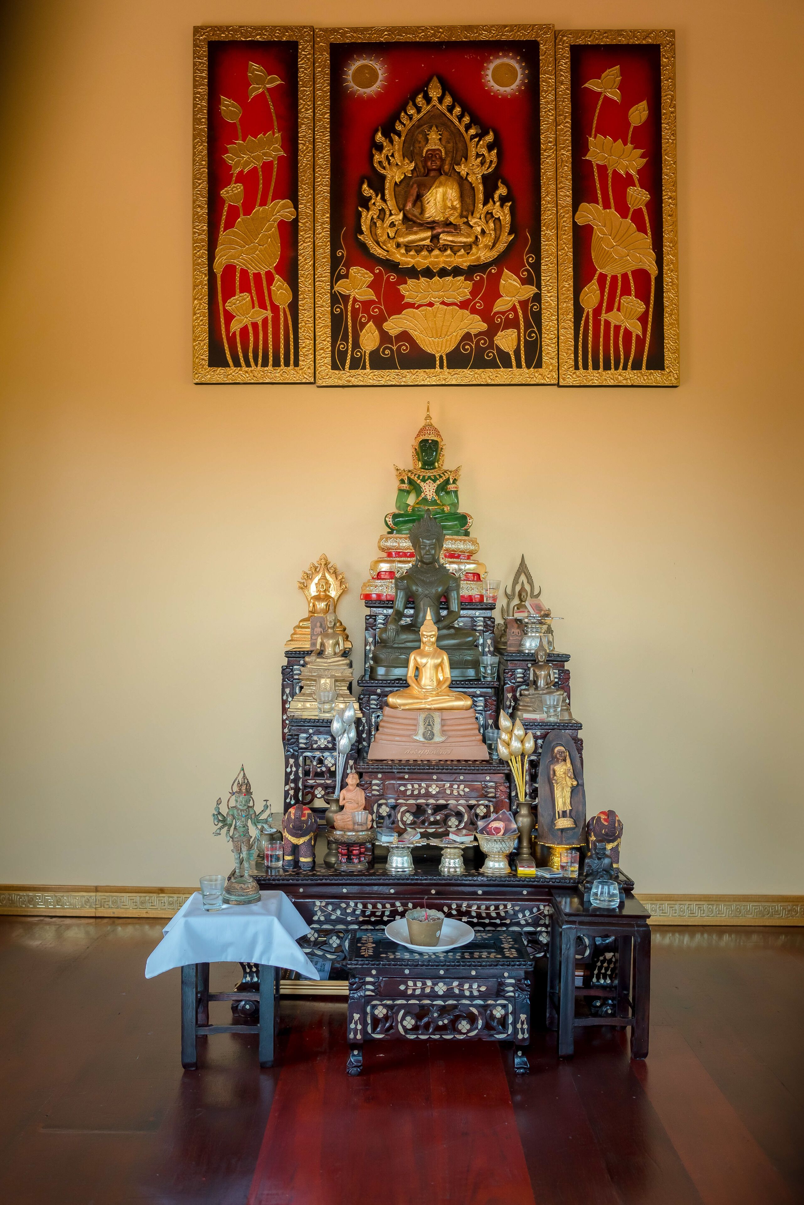 A room with a wooden altar featuring statues and religious artifacts, a white table with a glass, and a red and gold decorative panel above.