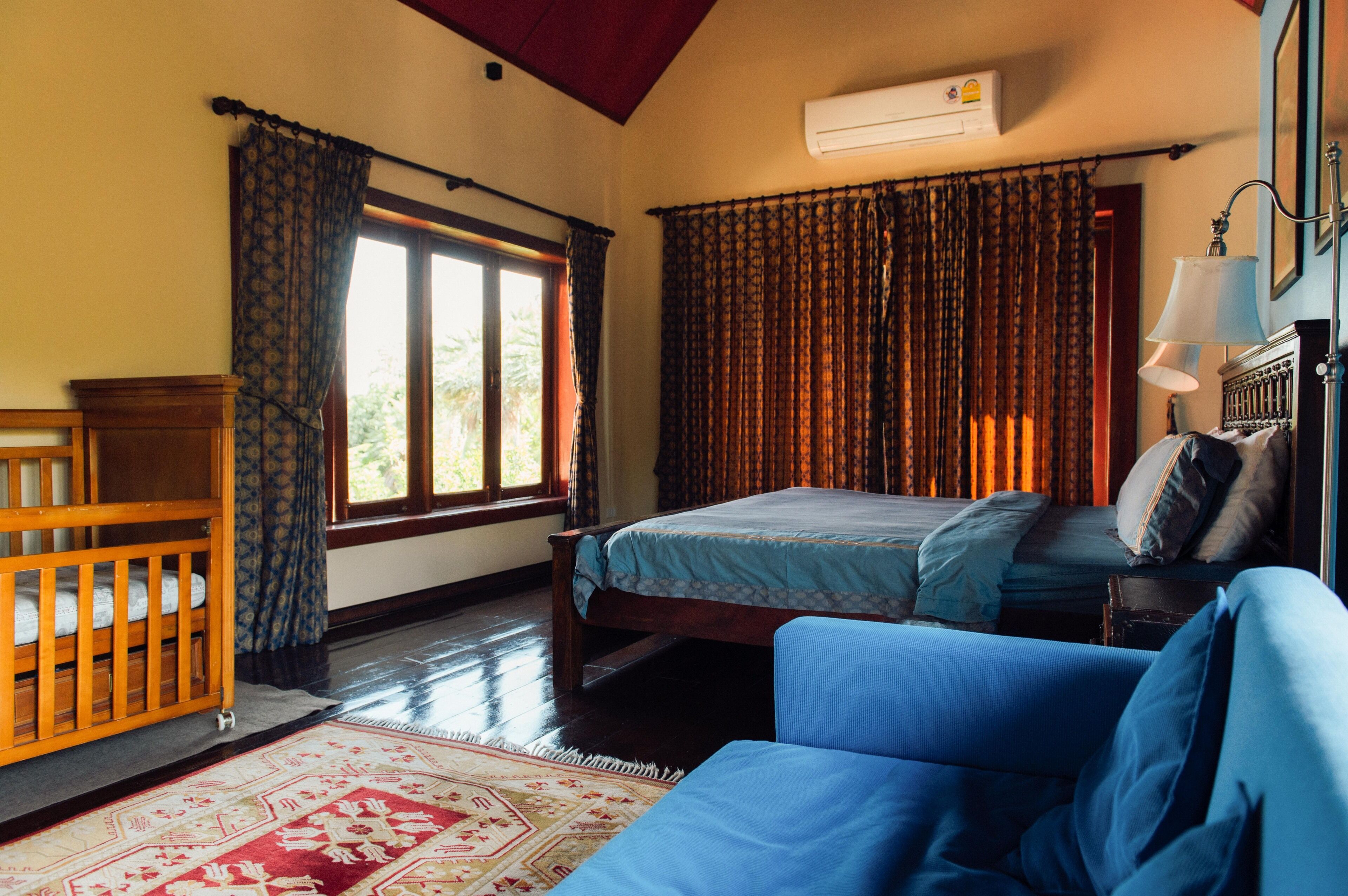 Un dormitorio con cama, cuna de madera, sofá azul, alfombra estampada y ventana con cortinas.