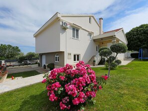 A two-story house with a white exterior, a visible air conditioning unit, a small front porch, and a well-maintained lawn with pink flowers.
