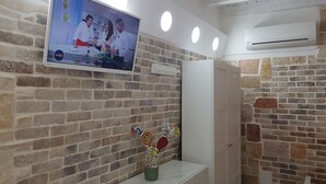 A reception area with a TV displaying a cooking show, a brick wall, a white door, and a small table with a decorative item. - New LOFT on Ortigia waterfront for max 3 guests (siracusa)