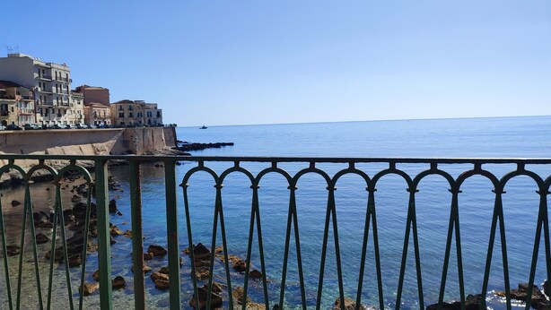 A coastal cityscape with buildings along the waterfront, a clear blue sky, and a metal railing in the foreground. - New LOFT on Ortigia waterfront for max 3 guests (siracusa)