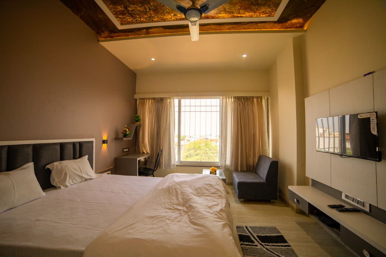 A bedroom with a large bed, a TV mounted on the wall, a desk, and a window with curtains.