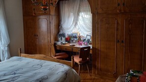 Ein Schlafzimmer mit einem Holzschrank, einem Bett mit floralem Überwurf, einem Holztisch mit Stuhl und einem großen Bogenfenster mit transparenten Vorhängen.