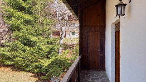 Eine Holztür mit steinbelagter Auffahrt, ein Balkon mit Holzgeländer und eine Landschaft mit Bäumen und einem Gebäude im Hintergrund.
