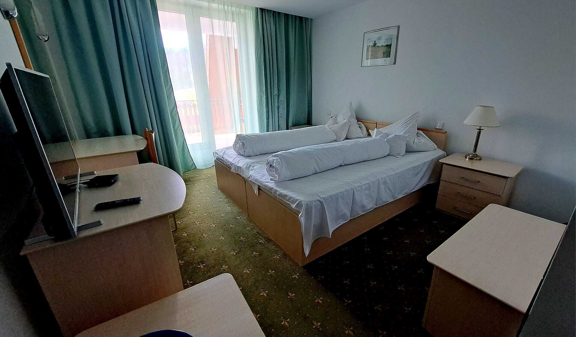 Habitación de hotel con cama, escritorio, silla, televisor, lámpara y ventana con cortinas.