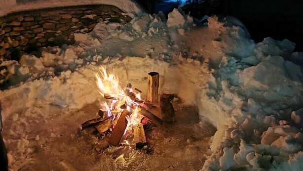 A fire burning in a snow-covered outdoor area with a stone wall and a building in the background. - Hotel Iadolina (Budureasa)