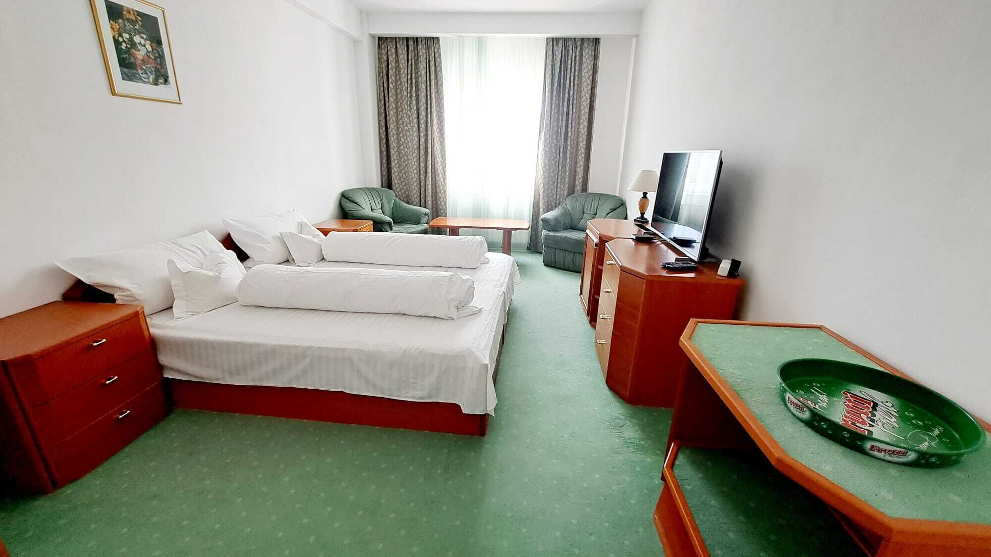 Habitación de hotel con cama, escritorio, silla, televisor y una mesita auxiliar verde.