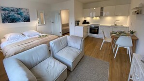 Une chambre d’hôtel moderne et compacte, comprenant un lit, deux canapés, un petit coin repas et une kitchenette.