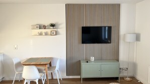 Un salon moderne comprenant un coin repas, un téléviseur fixé sur un mur à rayures, un meuble de couleur claire et un lampadaire.