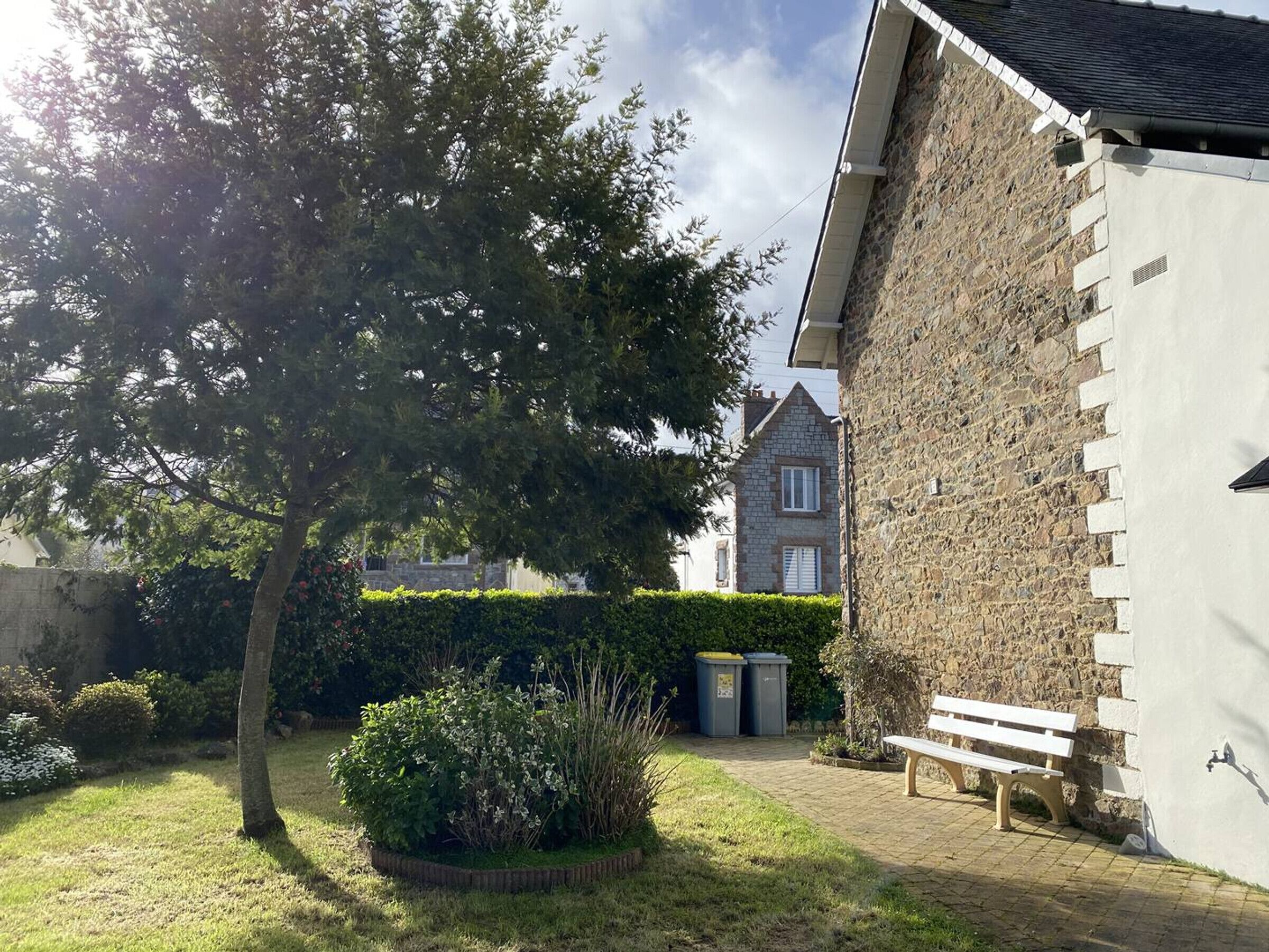 Un bâtiment en pierre à l’extérieur blanc, une cour pavée, un banc et un arbre.