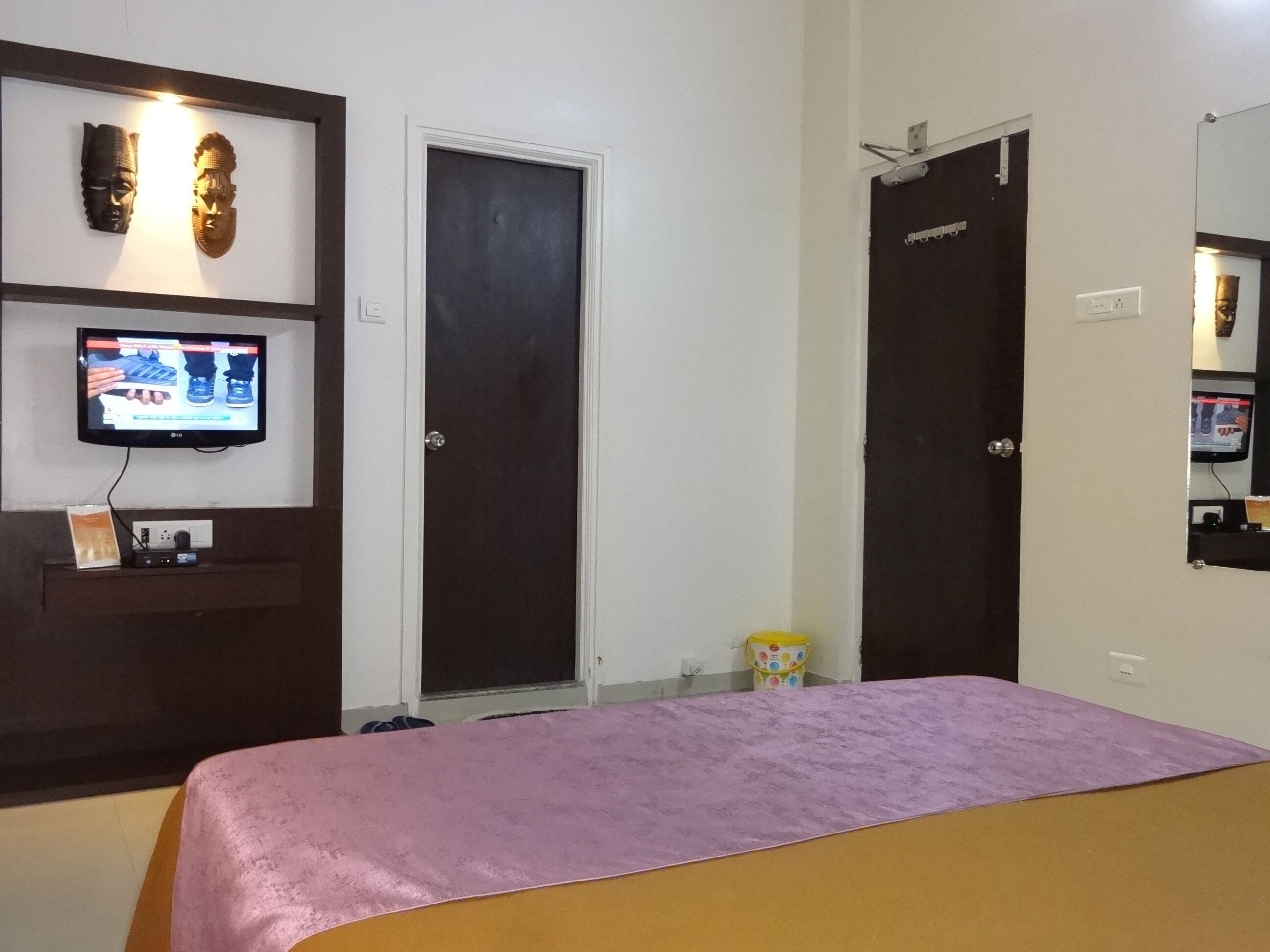 Un dormitorio con una cama, un televisor sobre un mueble, dos puertas y un cubo pequeño en el suelo.