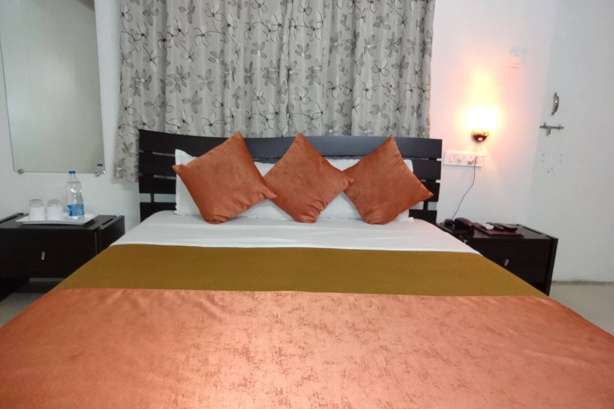 A neatly made bed with a brown bedspread and two orange pillows, a nightstand with a bottle and glasses, and a wall-mounted lamp.