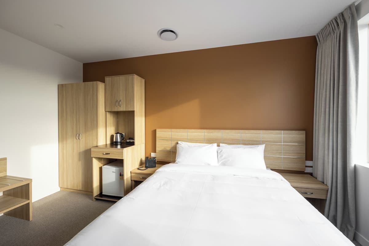 A hotel room with a bed, a wooden wardrobe, a small refrigerator, a bedside table, and a window with curtains.