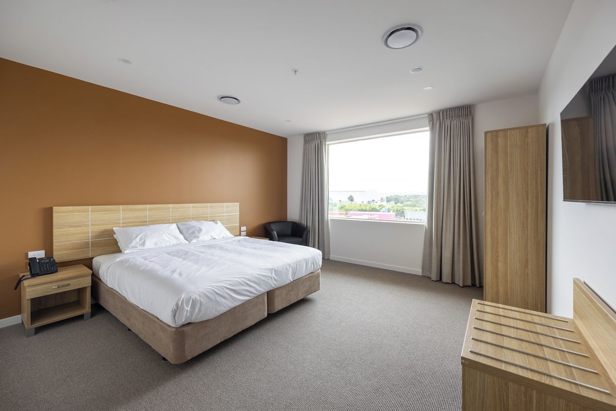 A hotel room with a bed, a nightstand, a desk, a chair, a wardrobe, and a large window offering a city view.