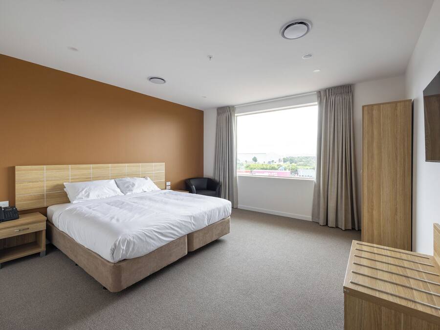 A hotel room with a bed, a nightstand, a desk, a chair, a wardrobe, and a large window offering a city view.