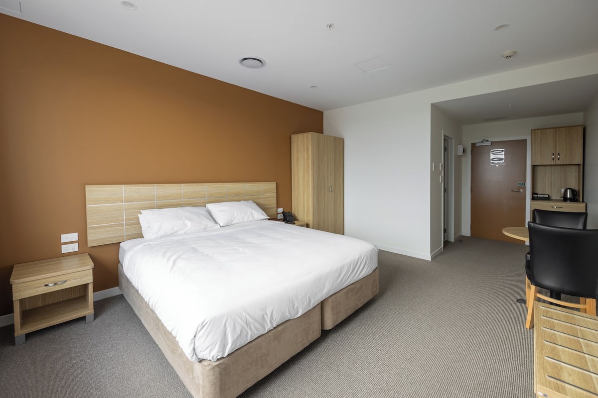 A hotel room with a large bed, a wooden wardrobe, a desk with a chair, and a kitchenette area.