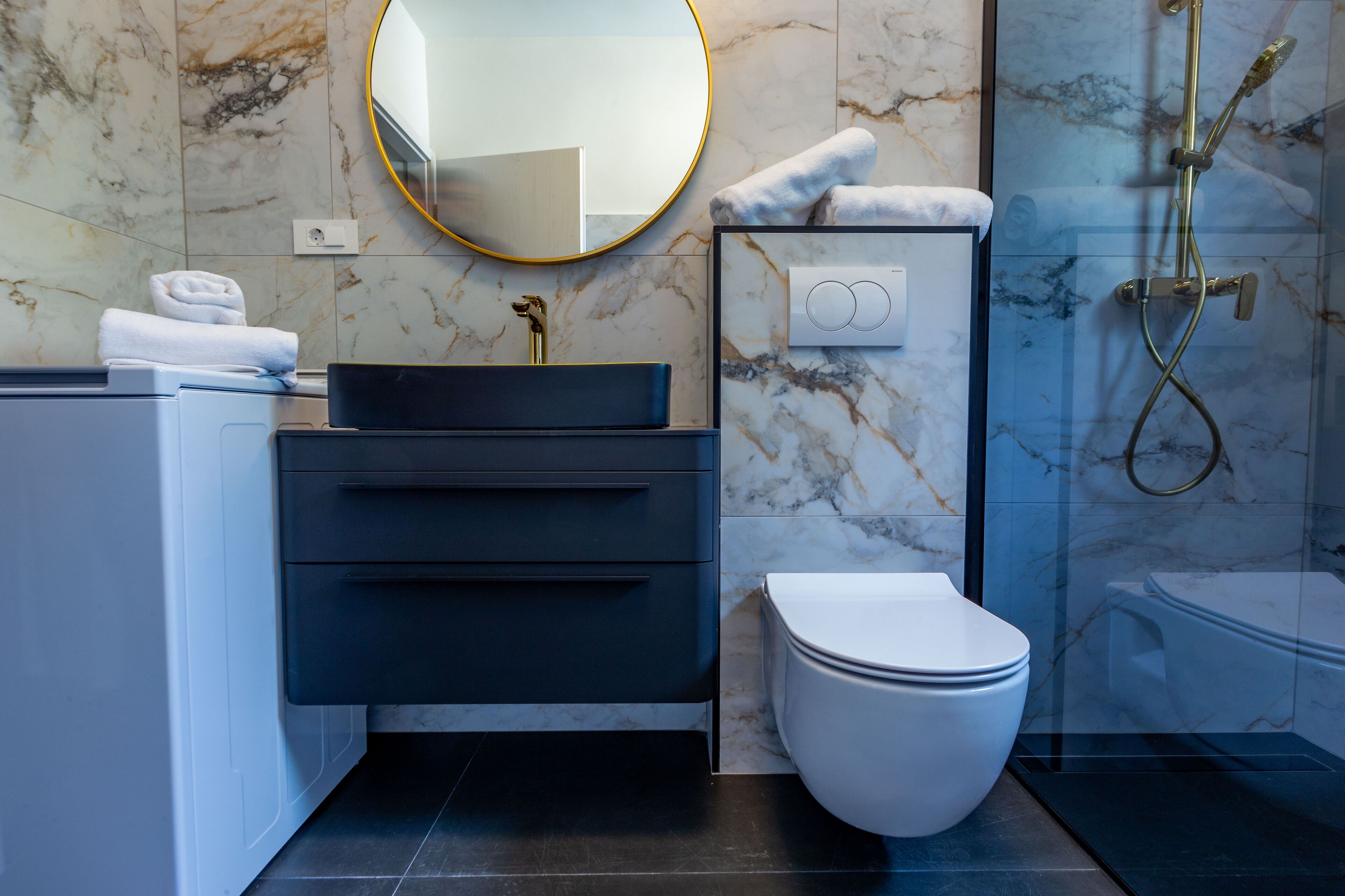 Een moderne badkamer met een marmeren wand, een ronde spiegel, een zwarte wastafel, een wit toilet en een douche met glazen wanden.