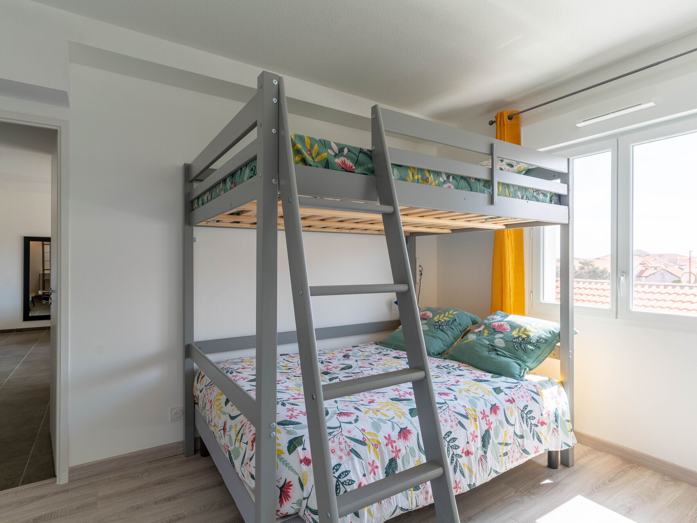 Uma cama beliche com escada, roupa de cama floral e uma janela com cortina amarela.