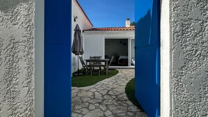 A stone pathway leading to a patio area with a table and chairs, a blue fence, and a building with a tiled roof. - 3-room villa 100m from the beach with patio and parking (La Palmyre)