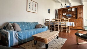 A living room with a blue-striped sofa, wooden coffee table, dining table with chairs, and a wooden cabinet with shelves. - 3-room villa 100m from the beach with patio and parking (La Palmyre)